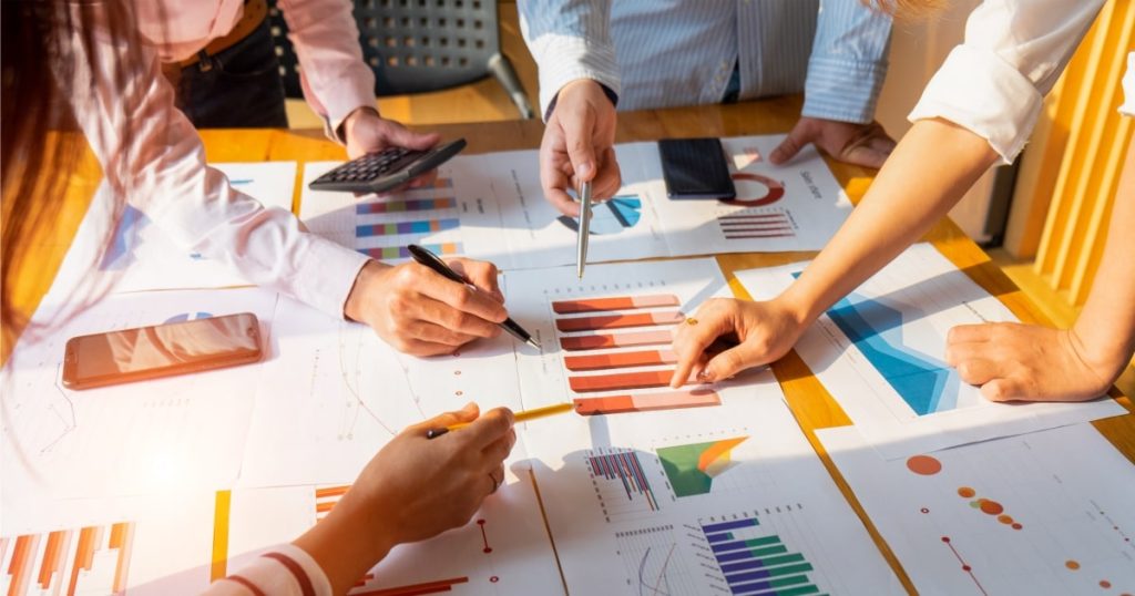 Manos de un grupo de personas señalando gráficos y datos en documentos sobre una mesa.