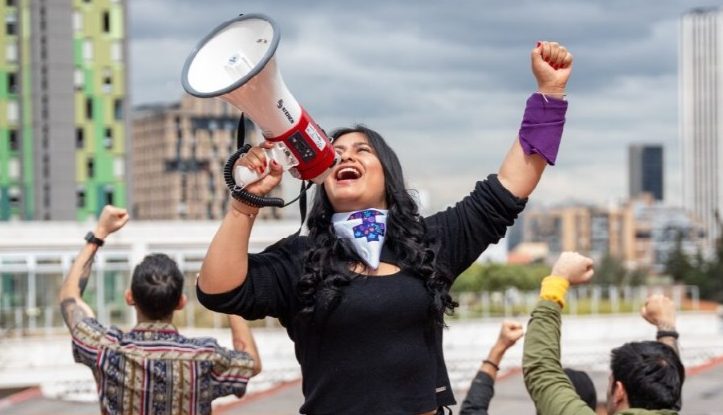 Mujer con pañuelo morado y megáfono alzando el puño durante una manifestación por los derechos de las mujeres.